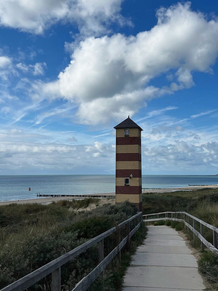 Ein Weg mit Holzgeländer führt durch die Dünen zu einem rot-weiß gestreiften Leuchtturm